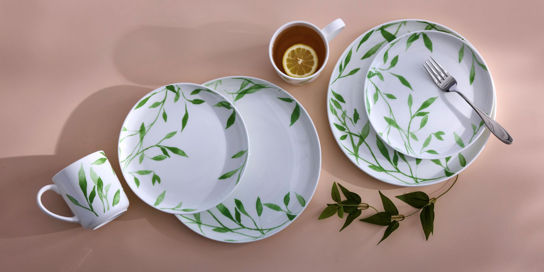 Akira collection white dinnerware with a green leafy pattern on a peach background, featuring plates, mug, lemon tea cup, and fork.