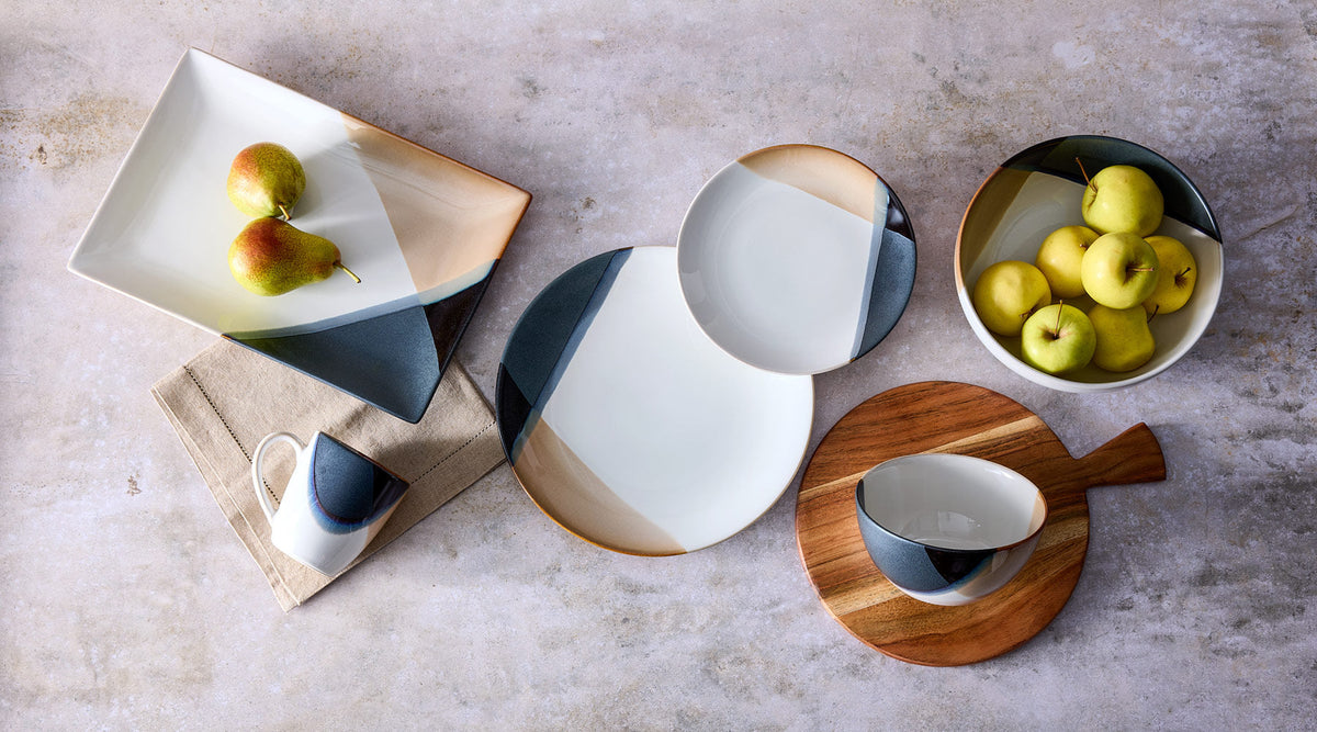 Caden ceramic dishware collection laid out on a textured stone surface with color block plates bowls a mug and a bowl of pears and apples.