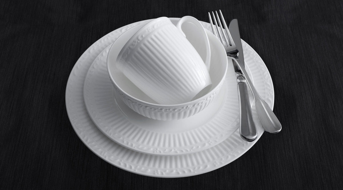 White bone china dinnerware stack with a ribbed cup inside a bowl atop nested plates and silverware on a dark surface.