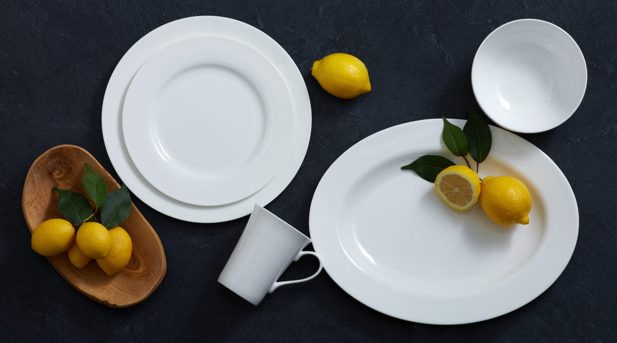 Delray collection flat lay of white dinnerware with plates, a mug, a bowl, and lemons with leaves on a dark surface.