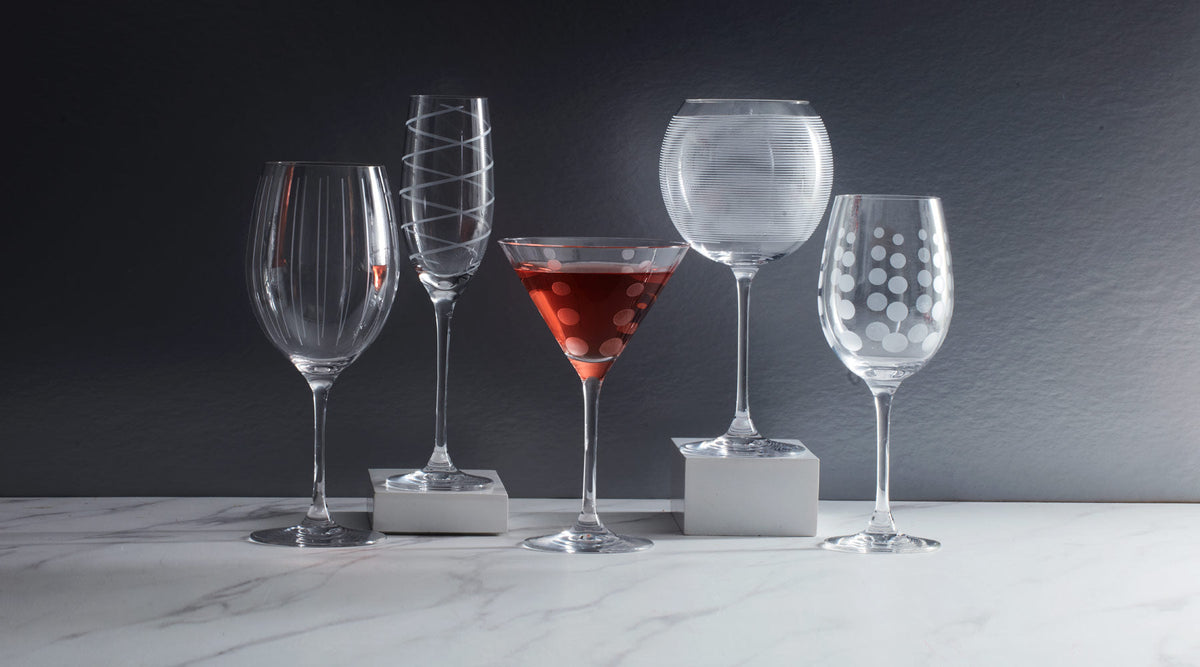 Assorted drinkware including an etched goblet, spiral-pattern flute, red dotted martini glass, ribbed round bowl glass, and polka dot wine glass on a marble surface.