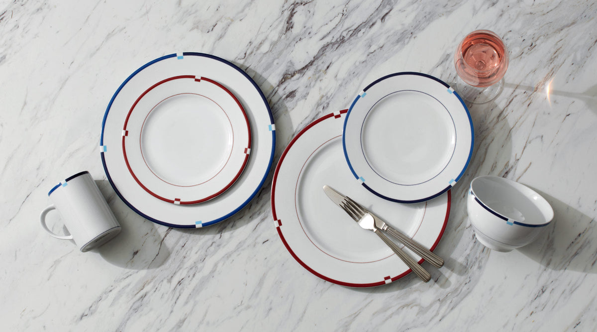 Jet Set dinnerware arranged on a marble tabletop with blue and red rimmed plates bowls a mug and a pink wine glass.