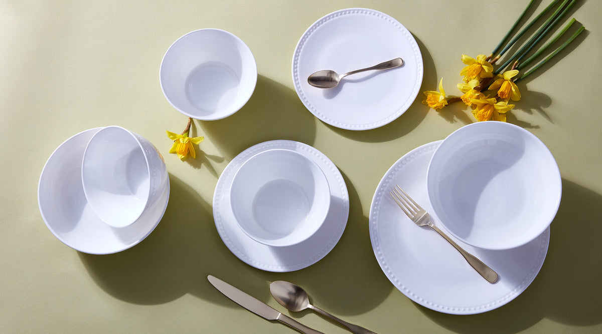 Overhead view of white dinnerware including plates and bowls with gold-toned flatware on a pale olive background, accented by yellow daffodils.