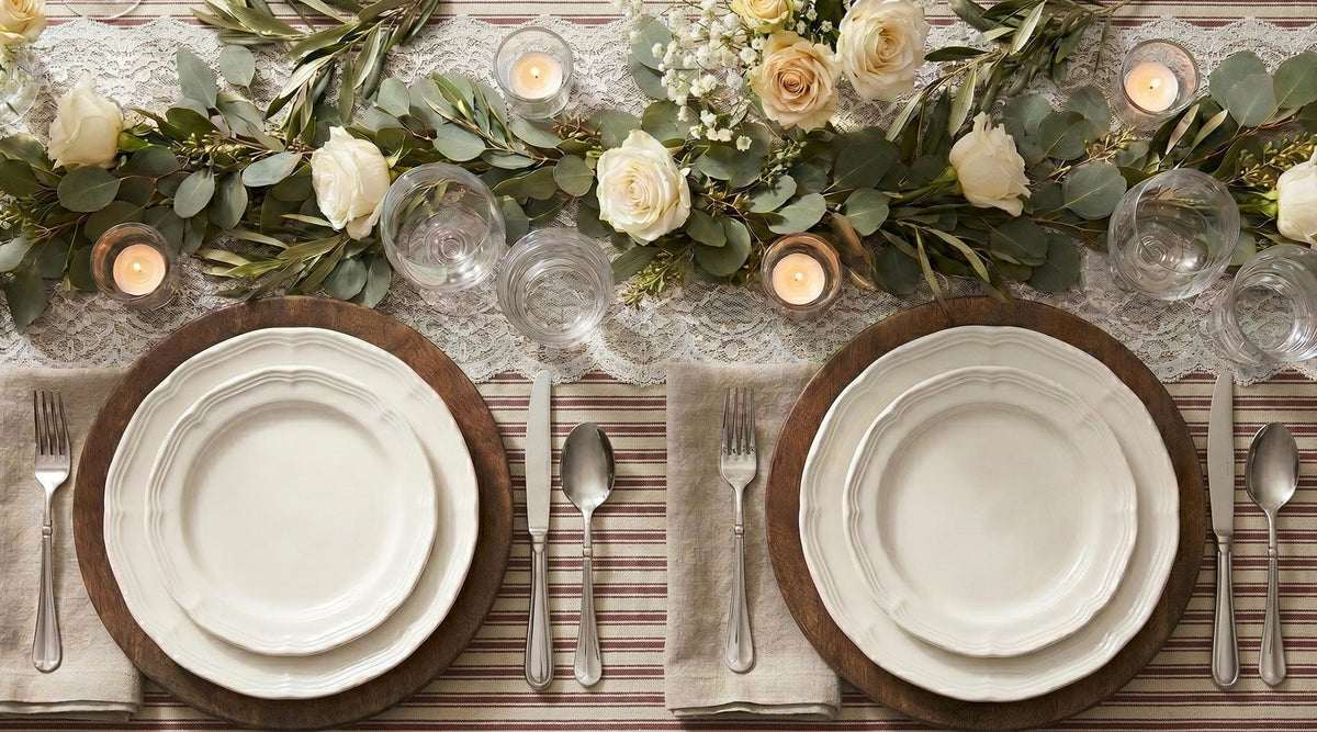 Rustic tablescape with white plates on wooden chargers, linen napkins, and a cream rose greenery garland with candles.