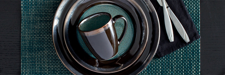 Top-down view of a teal placemat with stacked dark plates and a glossy mug, silverware on a black napkin.