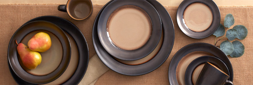 Sandstone dinnerware collection on a burlap surface with layered black and taupe plates and bowls, and a pear-filled bowl beside eucalyptus.