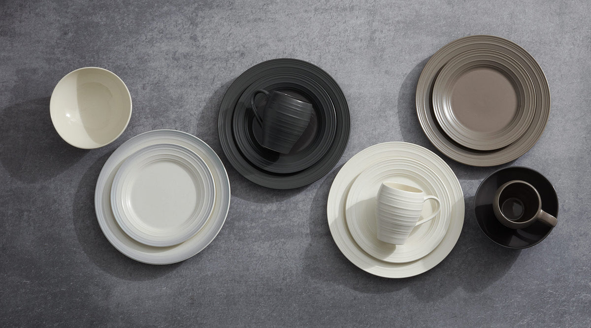 Swirl dinnerware collection on a gray surface with concentric ribbed plates in cream white gray and taupe plus mugs and a small beige bowl