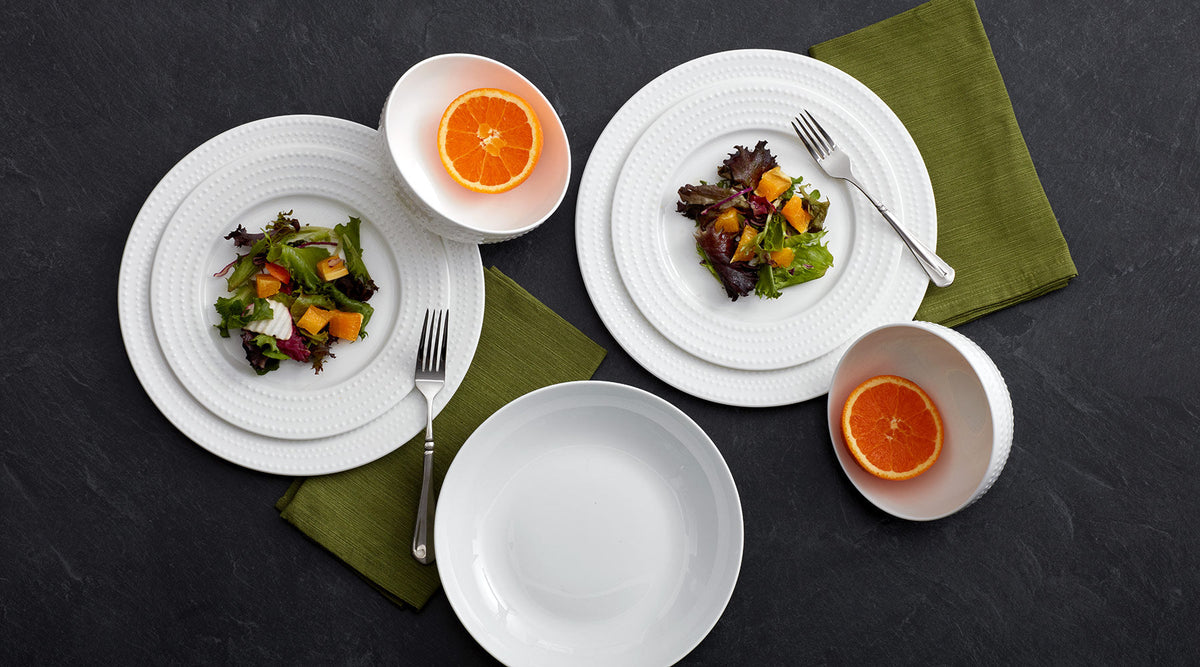 Overhead view of Nellie white beaded-edge dinnerware with salads, orange halves in bowls, and green napkins on a dark surface