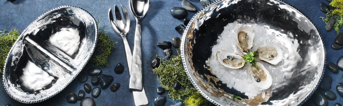 Two hammered Wilton Armetale serving bowls with cutlery on a blue surface, one bowl filled with oysters on ice.