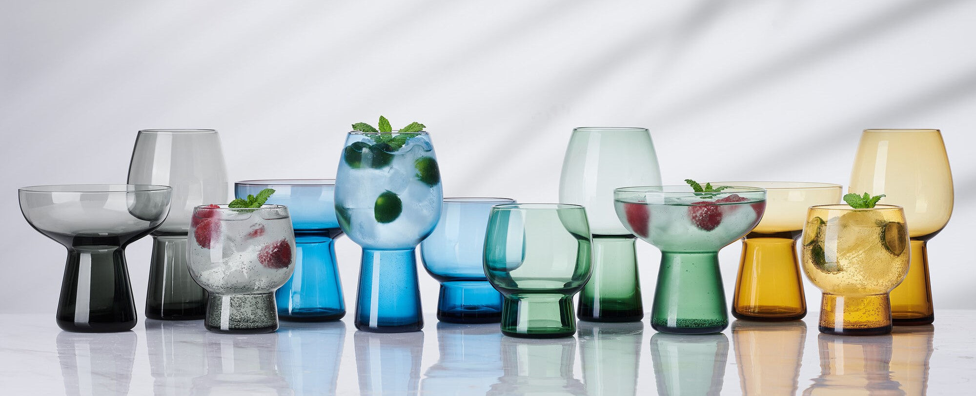 Phoebe glassware collection: row of colorful goblets, tumblers, and bowls in blue green and amber on a reflective surface, some with mint garnish.