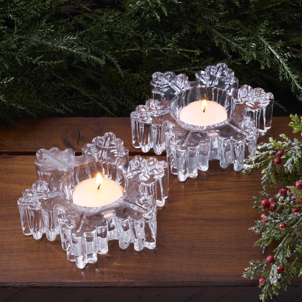 Two crystal snowflake tealight holders with burning candles on a wooden table surrounded by greenery.
