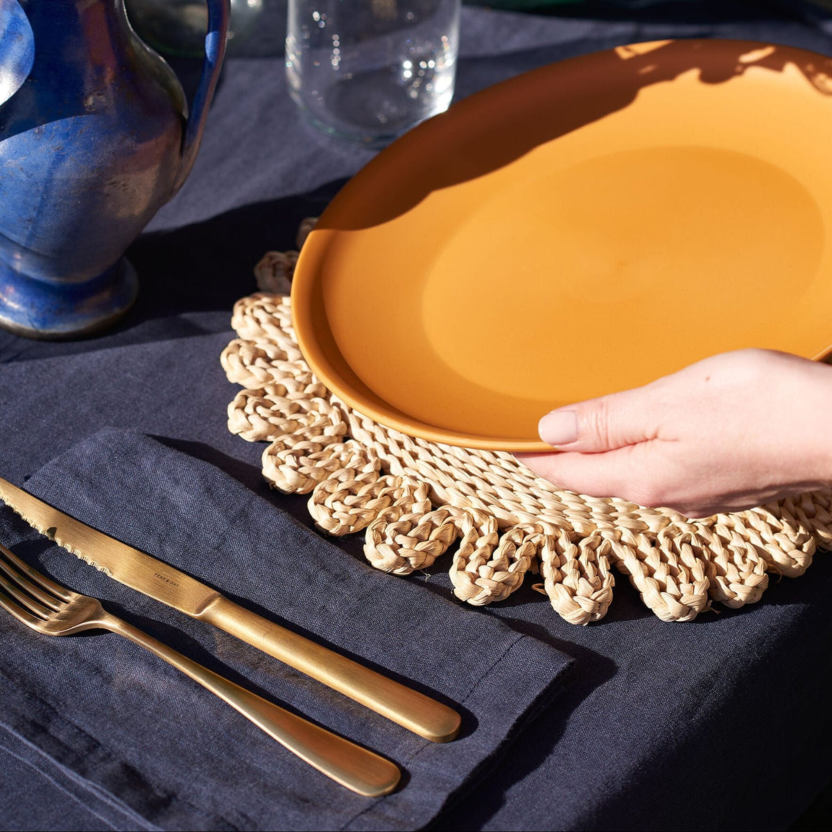 Midnight off-black linen tablecloth with a blue vase, clear glass and orange ceramic plate on a woven placemat.