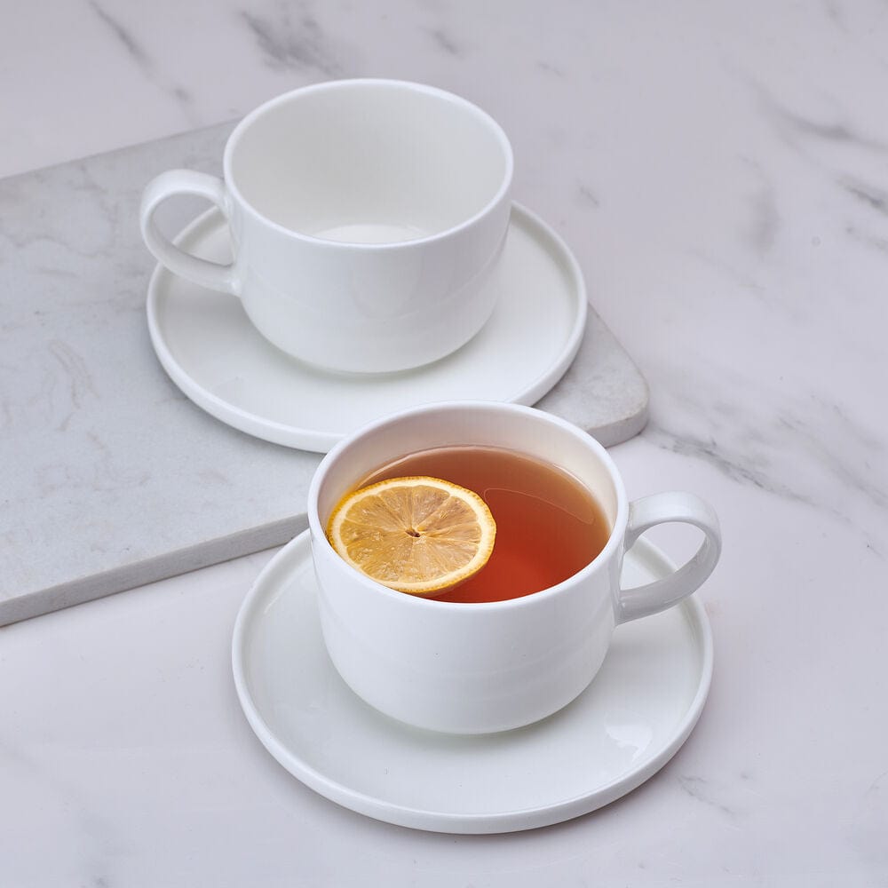 Two white teacups with matching saucers on a marble surface, one cup filled with tea and a lemon slice