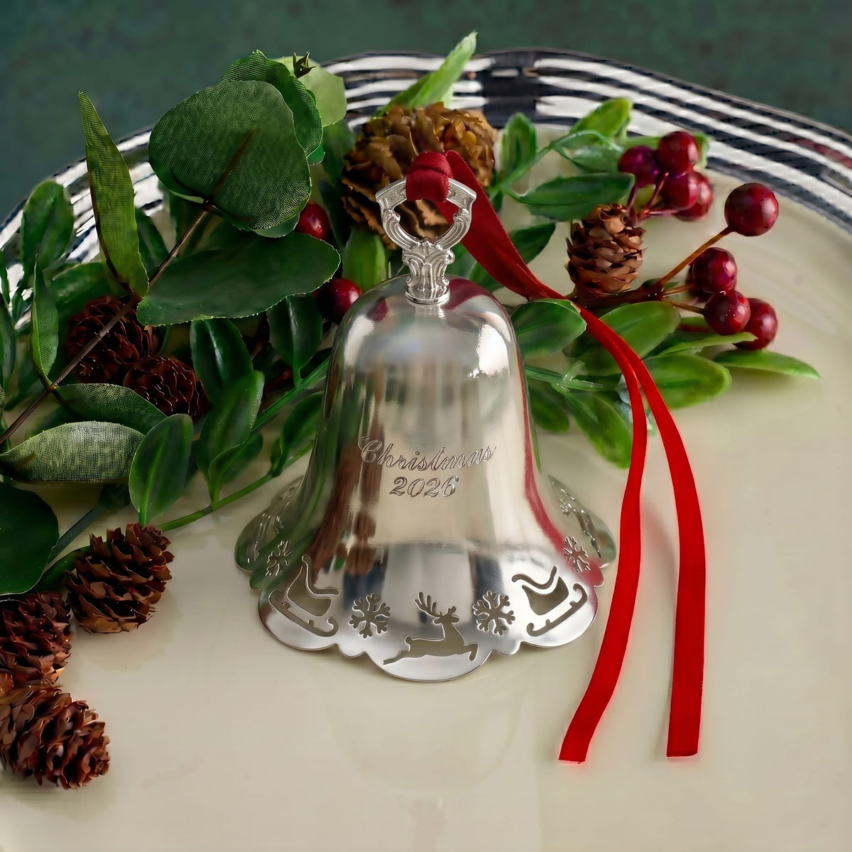 Silver pierced bell ornament nestled among greenery with a red ribbon and berry accents.