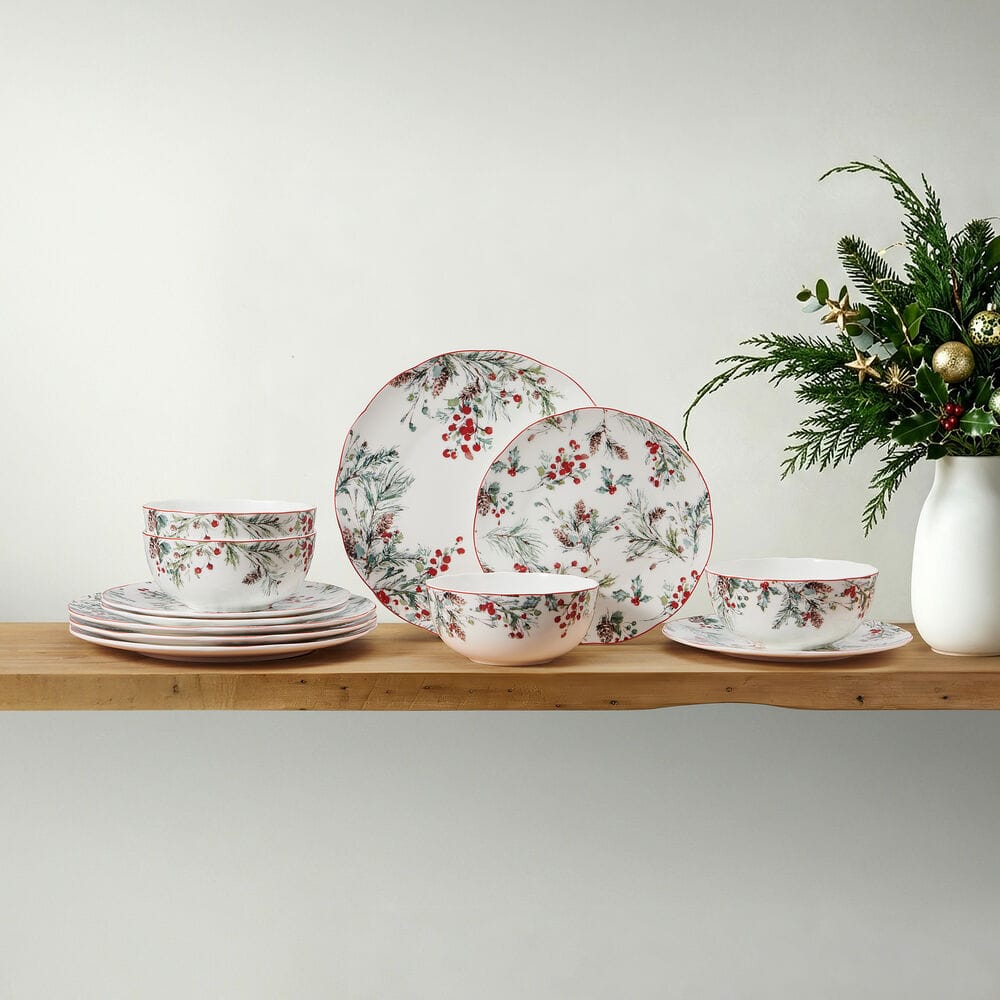 Pine and Berry dinnerware displayed on a wooden shelf beside a white vase with evergreen greenery.