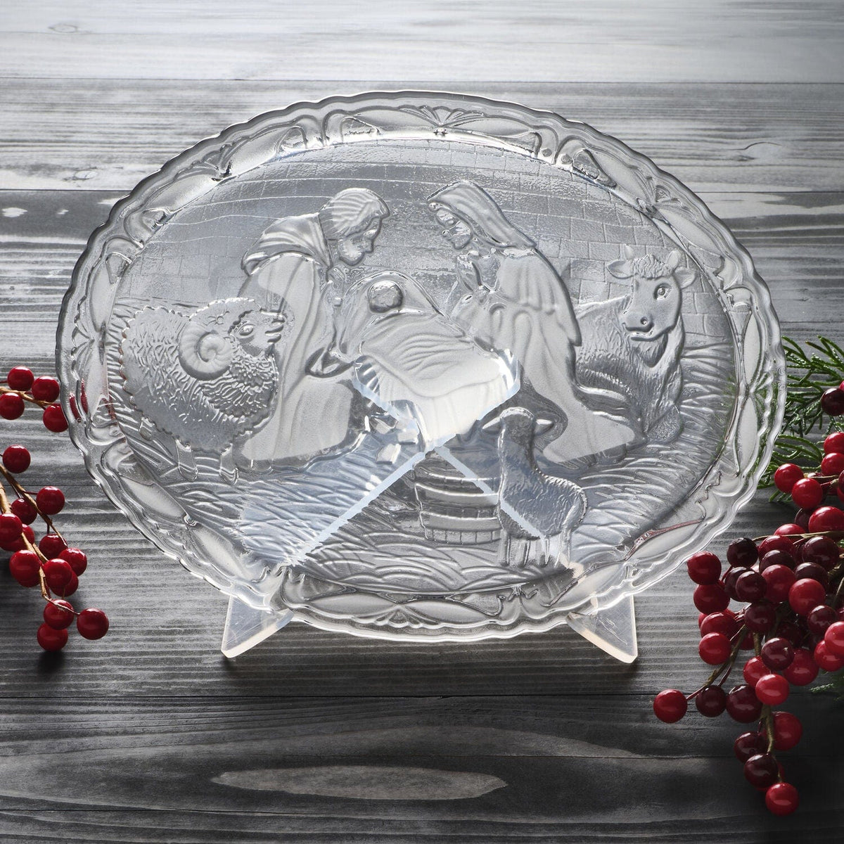 Crystal nativity plate with stand depicting the holy family and animals on a rustic wooden surface with red berries