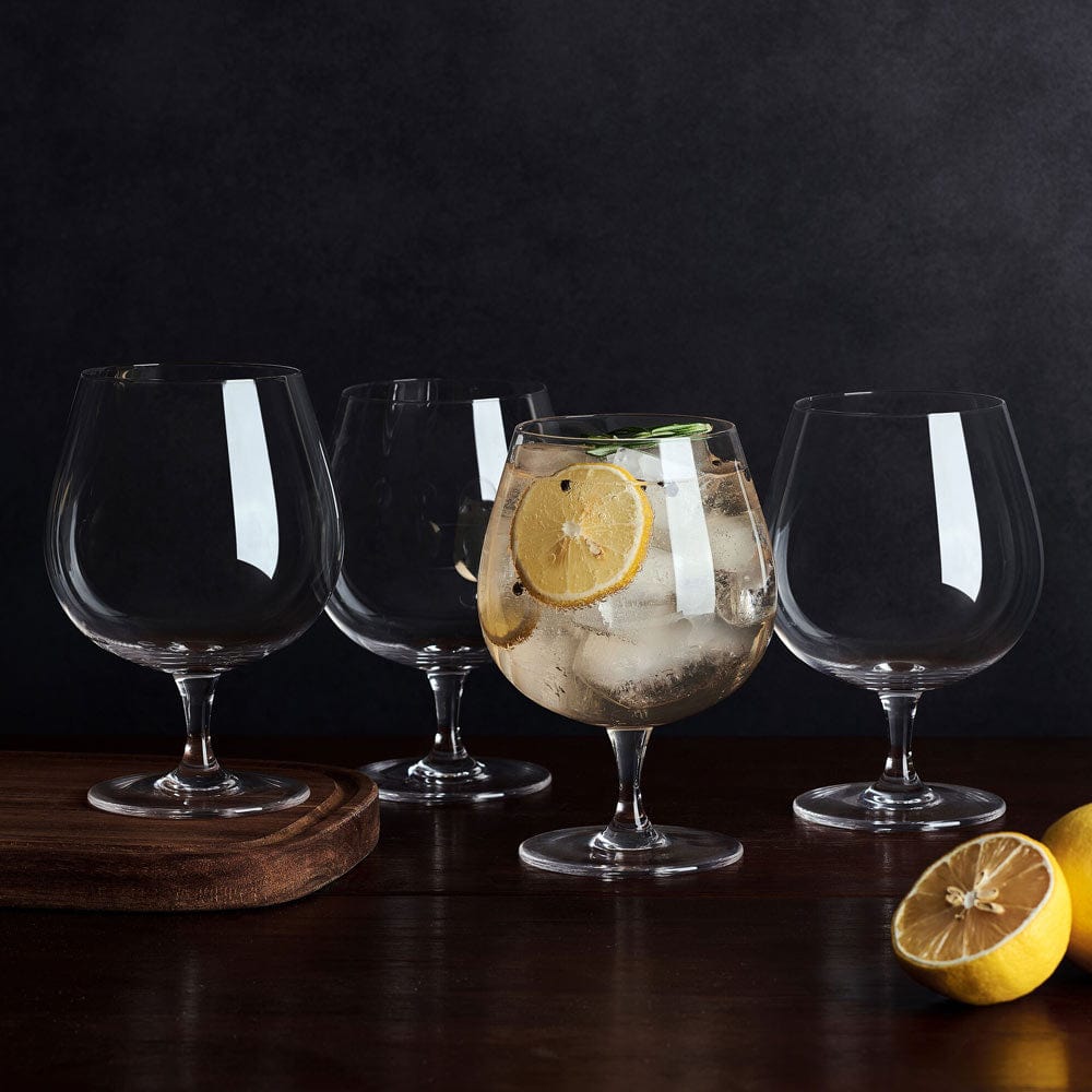 Four clear balloon glasses on a dark surface, one filled with a lemon slice and ice, with lemons nearby