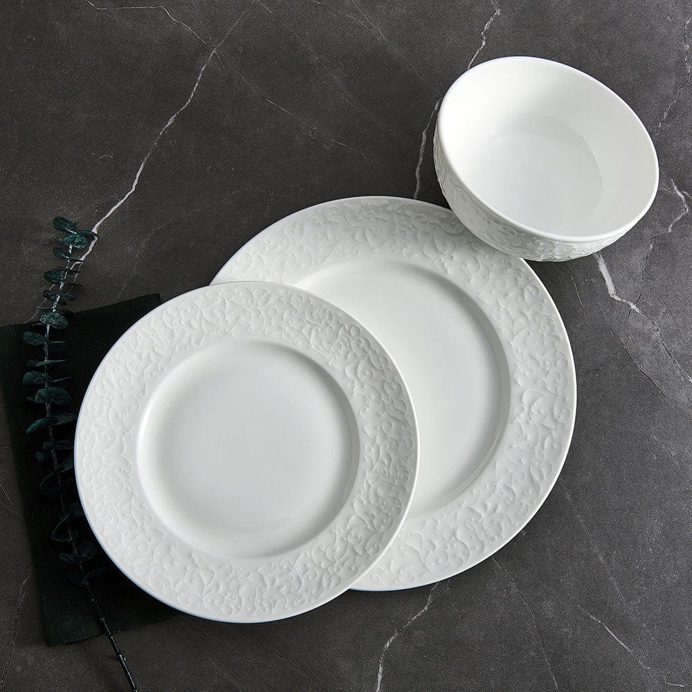 White embossed dinnerware set with floral border arranged on a dark surface including large plates and a bowl