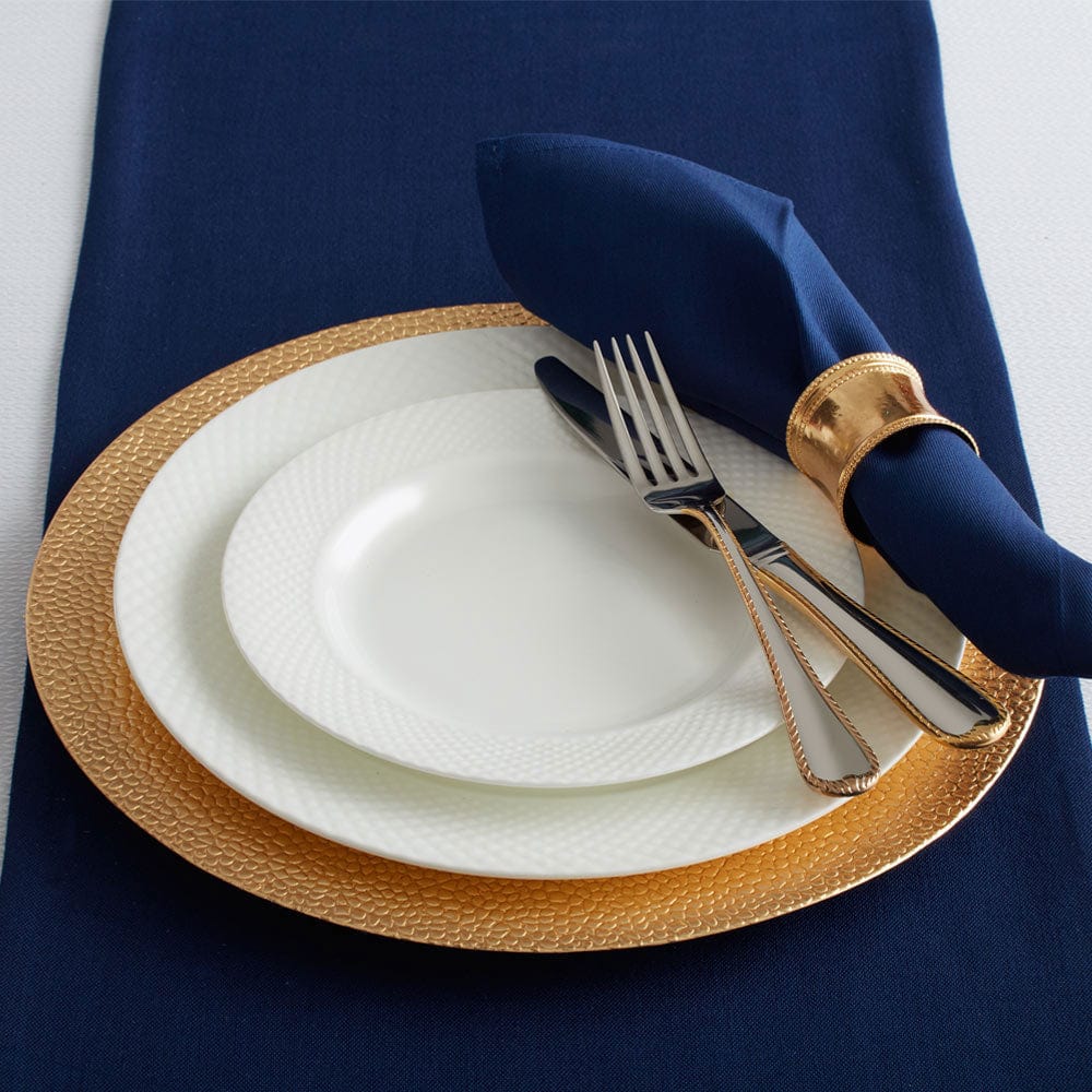 Navy blue table setting with a white Stanton dinner plate on a textured gold charger, silverware, and a navy napkin held by a gold napkin ring.