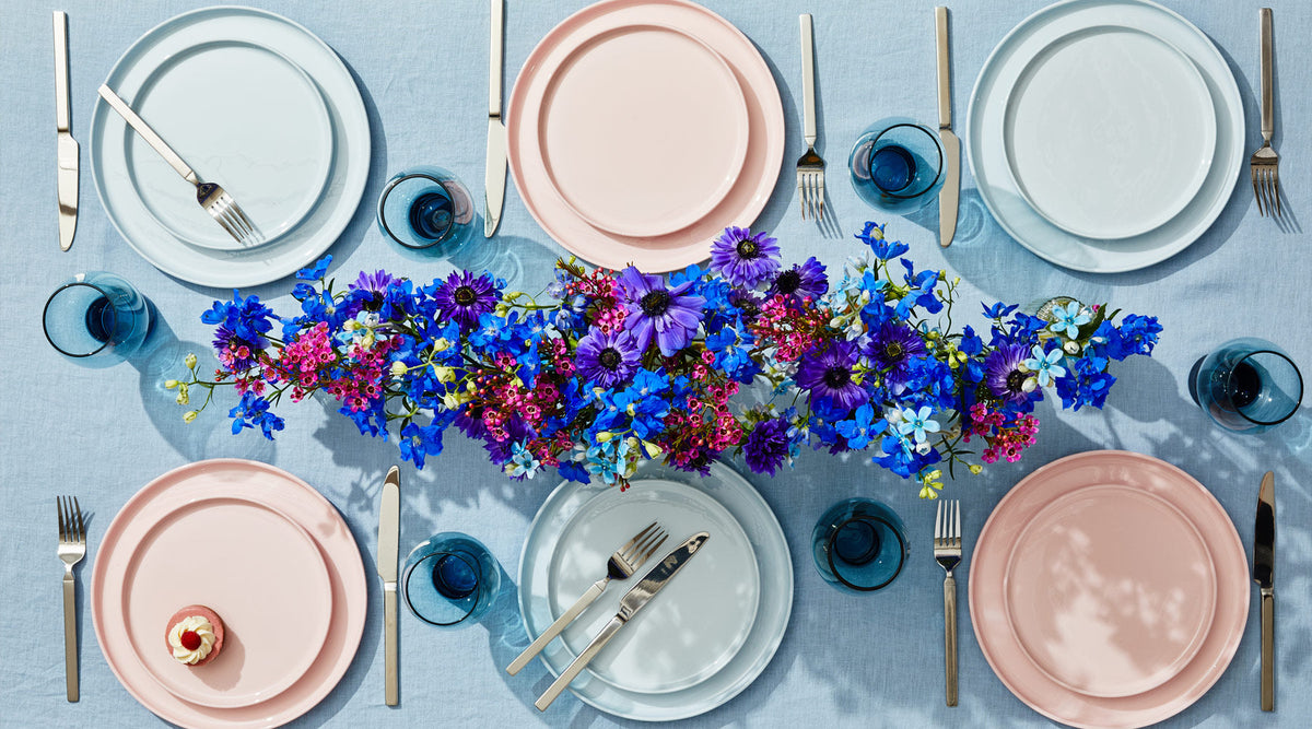 Pastel blue and pink bone china plates with silverware and blue glassware arranged around a vibrant flower centerpiece.