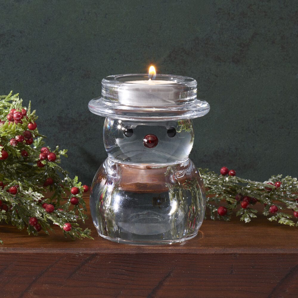 Clear glass snowman tealight holder on a wooden surface surrounded by festive greenery and red berries.