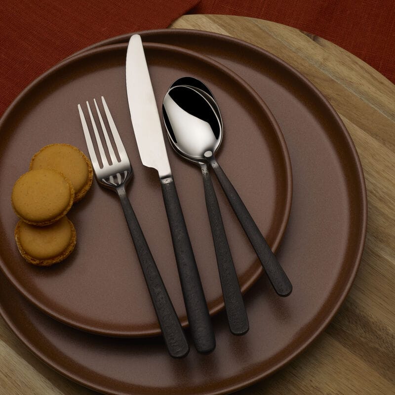 Calyx Black Forged flatware set laid on a brown plate with macarons nearby.
