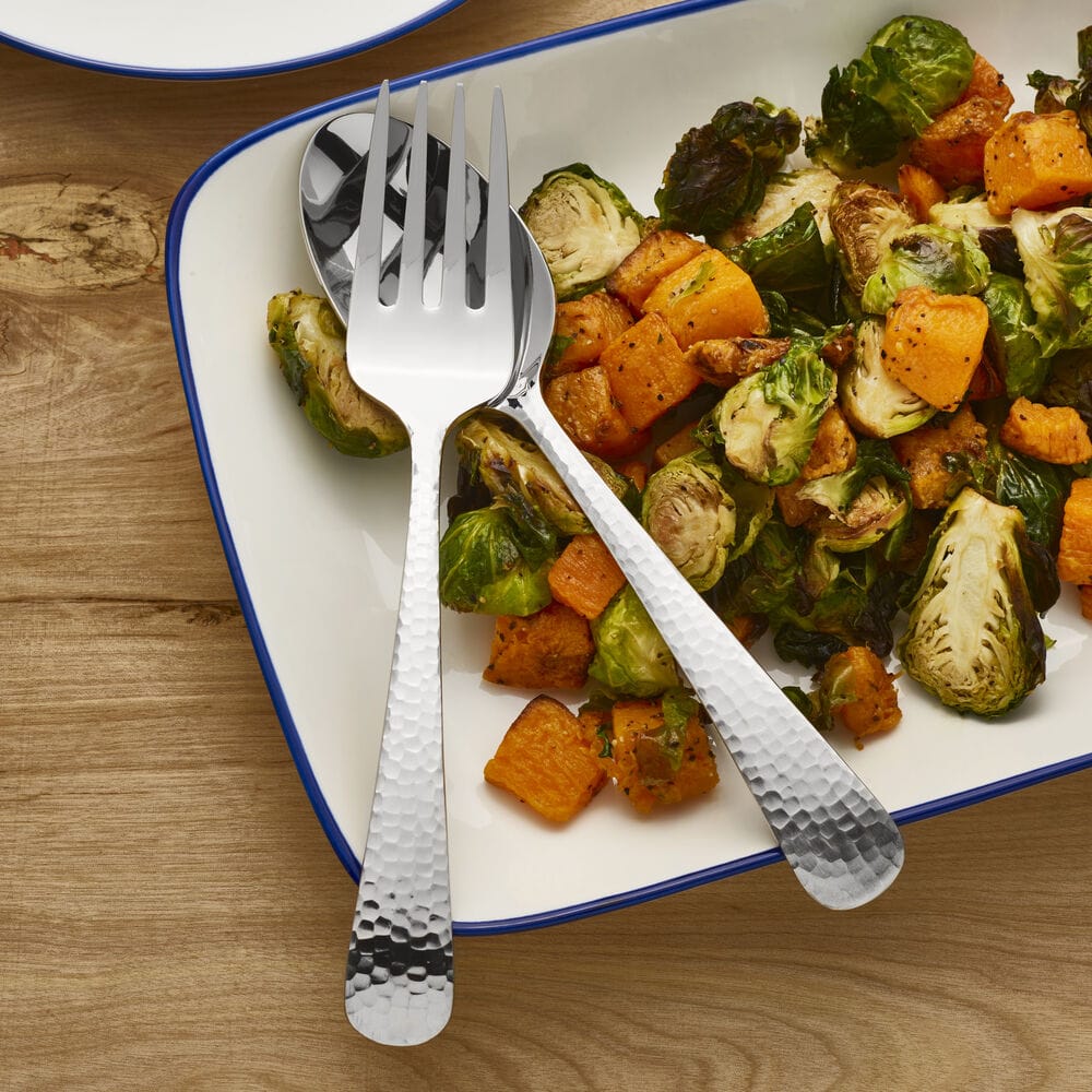 Roasted vegetables on a white plate with blue rim, hammered forks and spoons resting across.