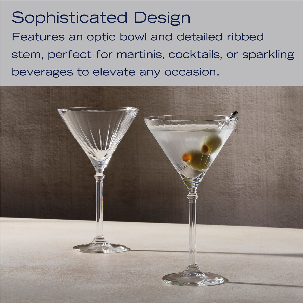 Two Eloise martini glasses with long stems on a light surface against a dark textured backdrop.
