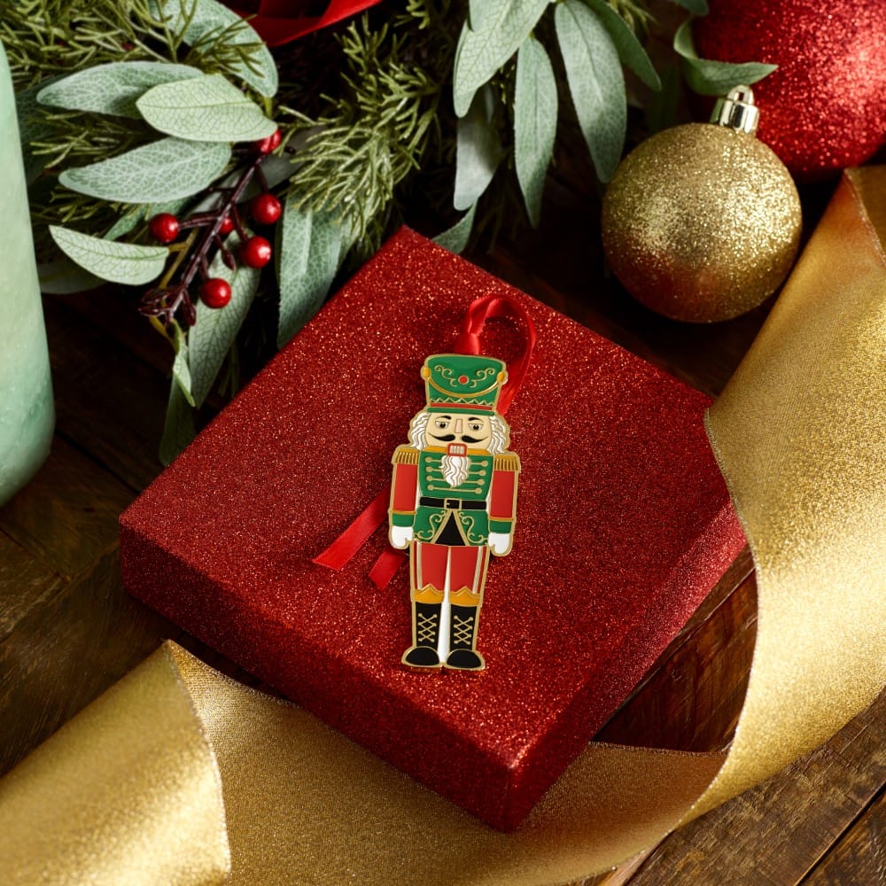 Nutcracker ornament resting on a glittery red gift box amid greenery and a gold ribbon