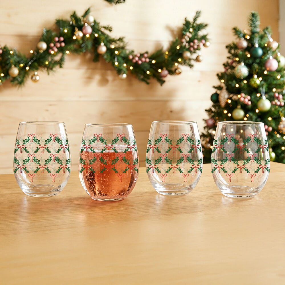 Set of four stemless wine glasses with green garlands and red bows on a wooden table, holiday decor in background.