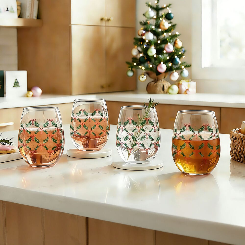Four stemless wine glasses with a holiday garland pattern on a kitchen counter with a Christmas tree in the background