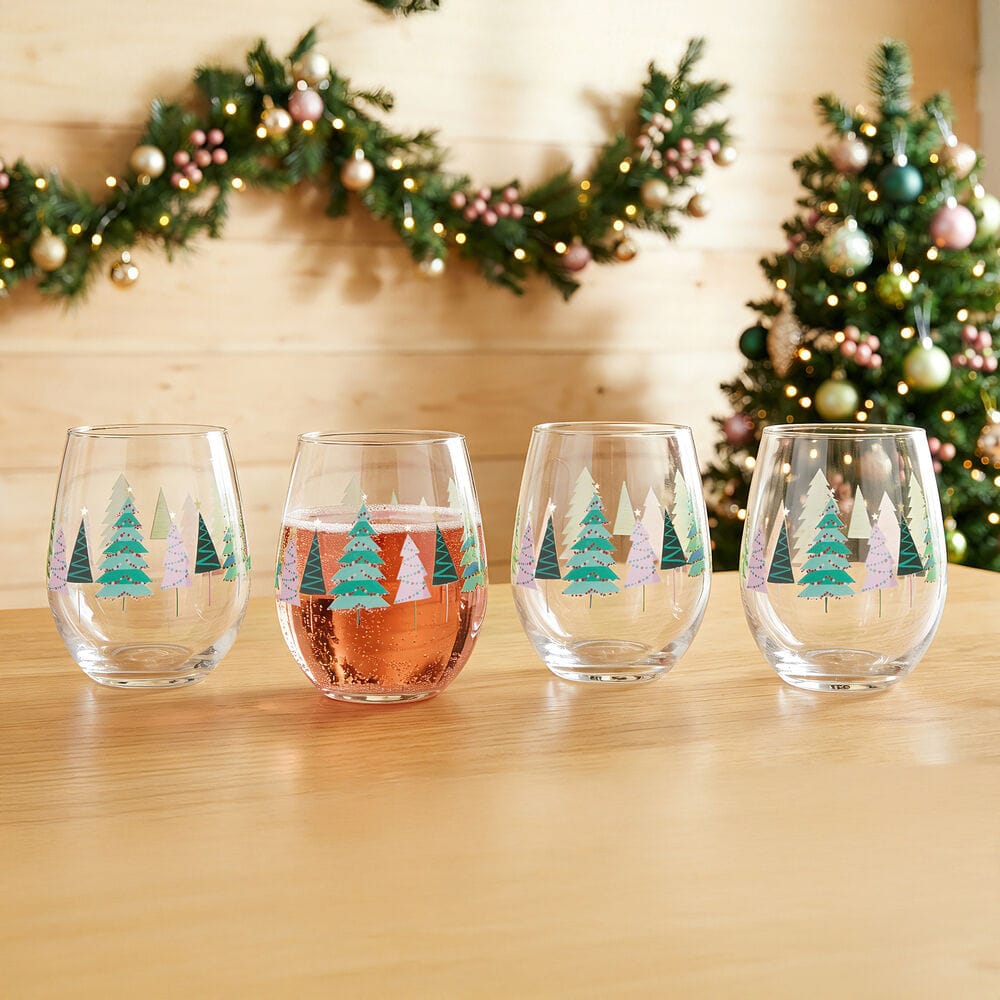 Four stemless wine glasses with colorful Christmas tree patterns on a wooden table with holiday garland in the background.