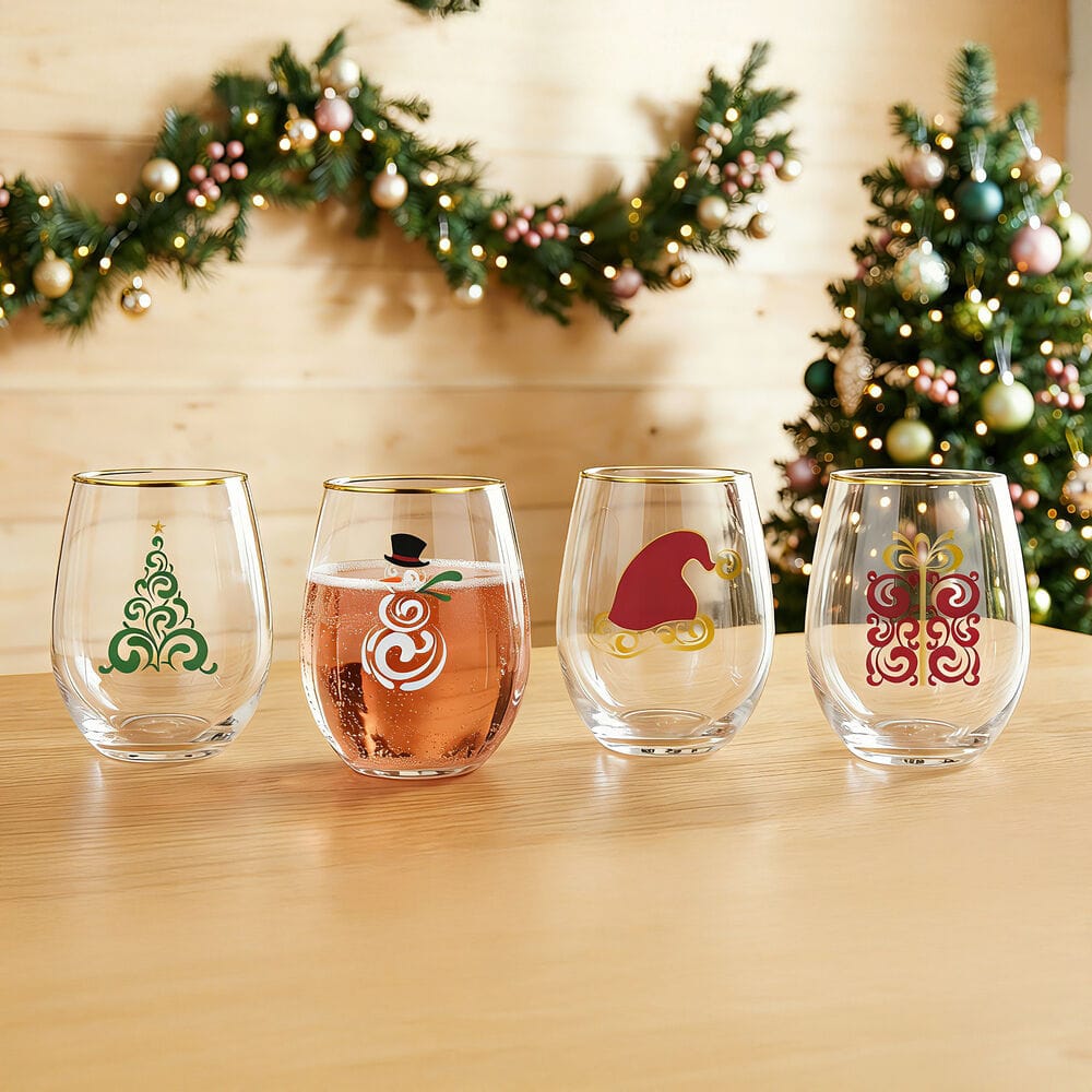Four clear stemless wine glasses with festive Christmas designs are lined up on a wooden table. The background features a decorated Christmas tree and a garland against a light wood wall.