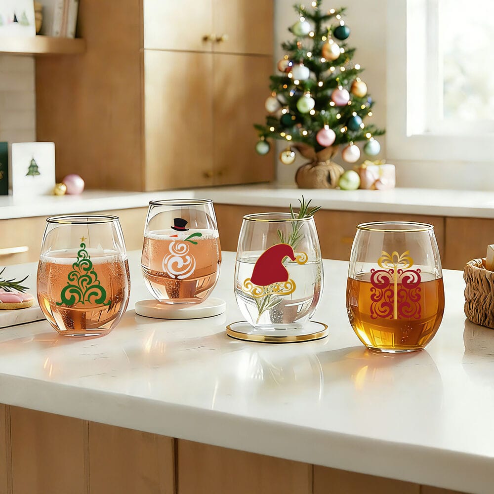 Four stemless wine glasses with festive Christmas designs are arranged on a white kitchen countertop. The background shows a decorated Christmas tree and wooden cabinets, indicating a holiday lifestyle setting.