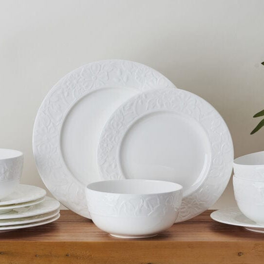 White Claire dinnerware set for four arranged on a wooden surface with embossed floral rims.