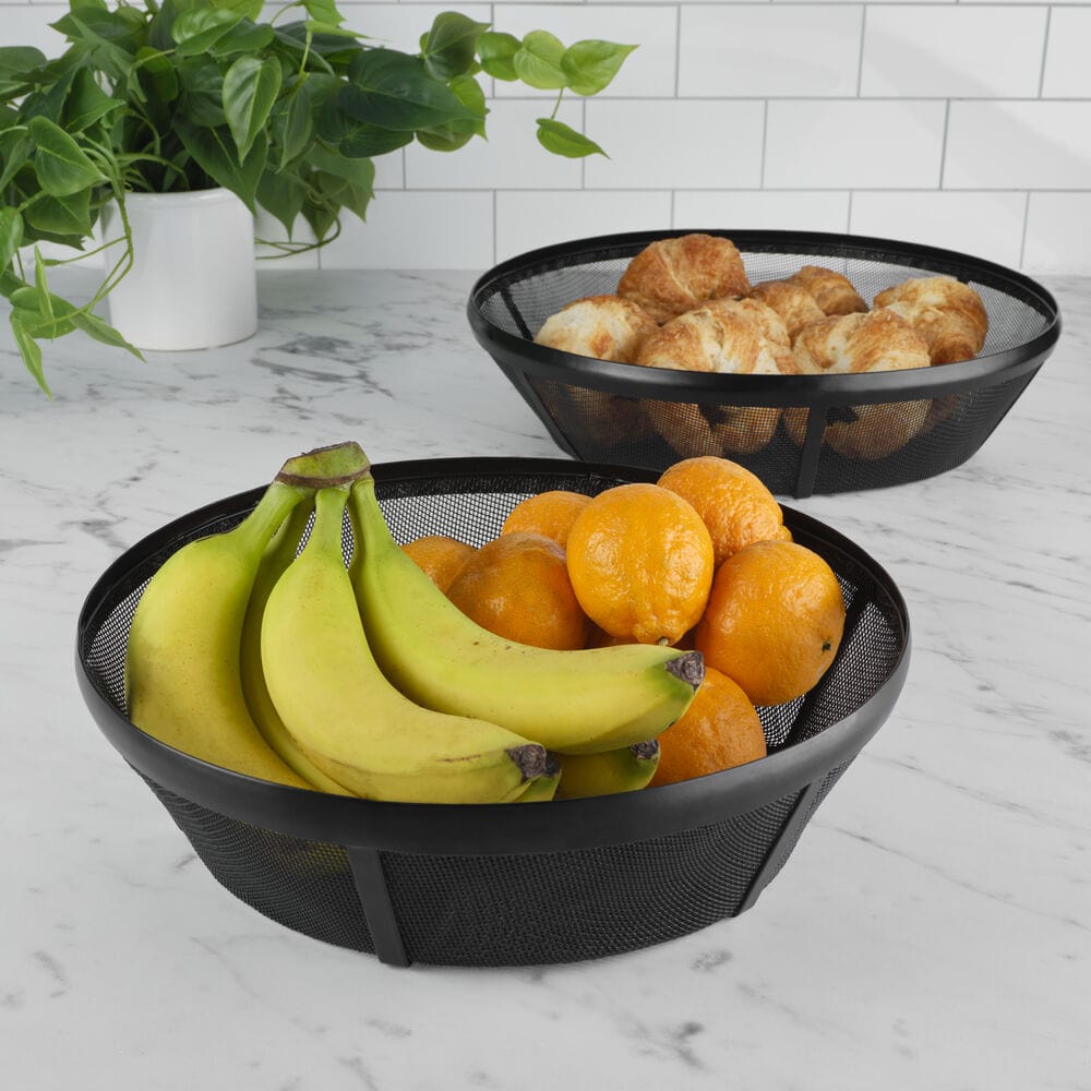 Two black mesh outdoor serving trays on a marble counter, one filled with bananas and oranges, the other with croissants.