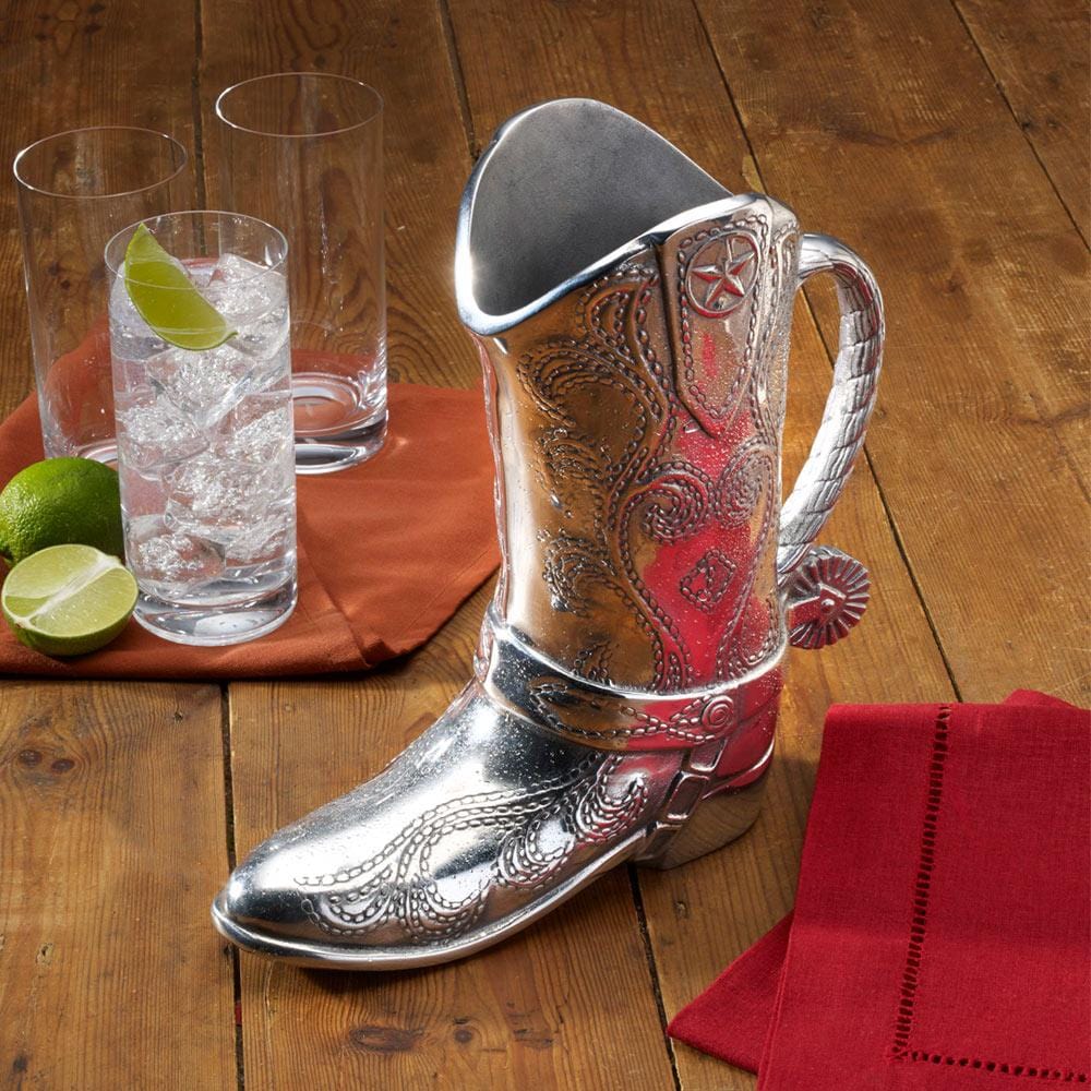 Silver cowboy boot pitcher set on a wooden table beside glassware and limes.