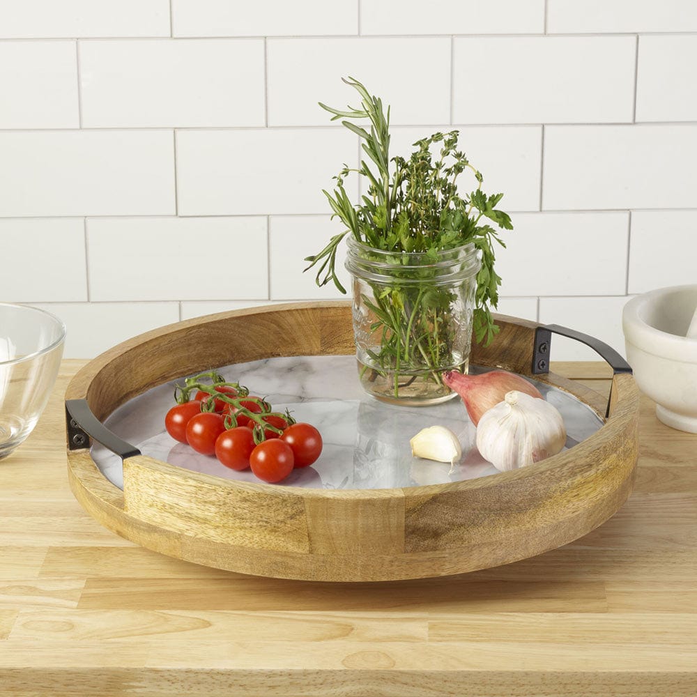 Round marble Lazy Susan with wood rim and black handles on a kitchen counter, holding a jar of herbs, tomatoes and garlic.