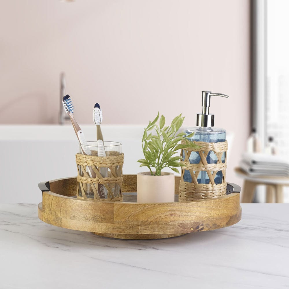 Round marble Lazy Susan with wood rim and black handles on a light counter, featuring a glass toothbrush holder, a small potted plant and a twine wrapped soap dispenser.